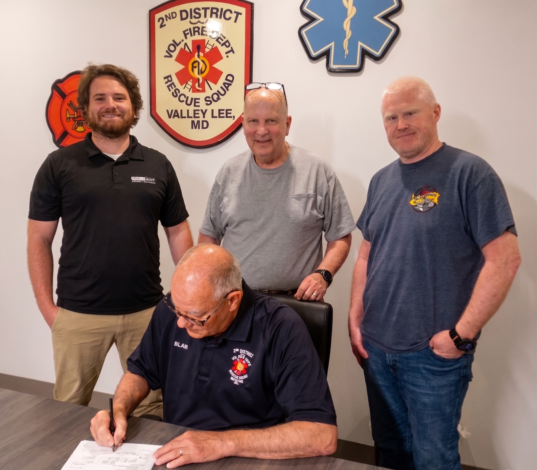 SDVFDRS President Blair Swann sits at a conference table signing the commercial solar contract. Standing behind him, from left to right, are Energy Select's Gregg Havens, SDVFDRS Solar Project Manager Jay Rodgers, and SDVFDRS Treasurer Mike Scrivener. Second District Volunteer Fire Department and Rescue Squad logos hang on the wall behind them.